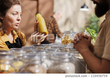 Vendor showcasing her products in zero waste marketplace venue, selling homemade fruit juice to interested customer. Local farmer trading environmentally conscious healthy food items during event 110456759