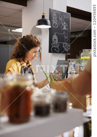 Storekeeper at zero waste supermarket checkout counter using bar code scanner to process customer transaction. Seller in sustainable local neighborhood shop scanning client food items 110456893