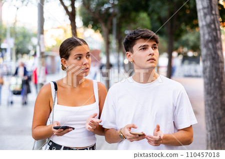 Young guy and girl using mobile phones while walking along summer city street 110457018