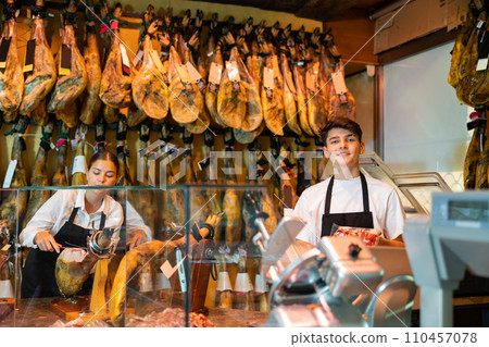 Girl and guy sellers of butcher shop working behind counter with Iberian jamon Girl and guy sellers of butcher shop working behind counter with Iberian jamon 110457078