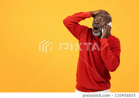 Shocked senior Black man in a red sweater holding a phone to his ear, wide-eyed 110458855