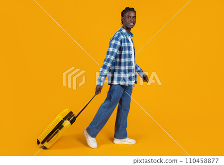 Smiling young black man casually walking with yellow suitcase on orange background 110458877