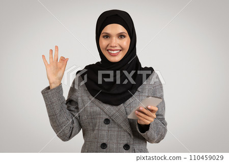 Cheerful woman in hijab, okay sign while holding phone 110459029