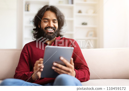 Relaxed eastern guy scrolling, using digital tablet at home 110459172