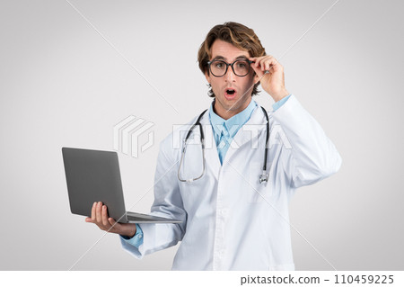Surprised male doctor with glasses holding laptop Surprised male doctor with glasses holding laptop 110459225