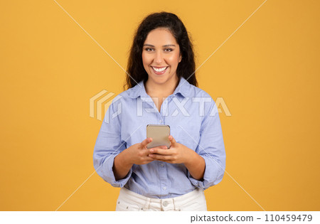 Happy young woman in a blue shirt holding a smartphone and looking at the camera Happy young woman in a blue shirt holding a smartphone and looking at the camera 110459479