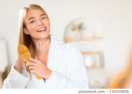 Blonde young lady in bathrobe combs her long hair indoors Blonde young lady in bathrobe combs her long hair indoors 110459606