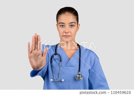 Determined european nurse in blue scrubs making a stop sign with her hand, a gesture of caution in healthcare 110459628