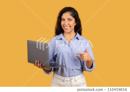 Friendly young woman with curly hair holds a laptop and points to it with a smile Friendly young woman with curly hair holds a laptop and points to it with a smile 110459658