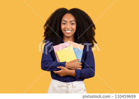 Happy african american lady student smiling at camera, holding copybooks, set against yellow background Happy african american lady student smiling at camera, holding copybooks, set against yellow background 110459689