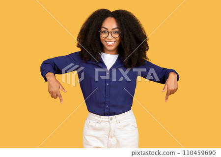 Smiling black woman pointing down, standing on yellow background Smiling black woman pointing down, standing on yellow background 110459690