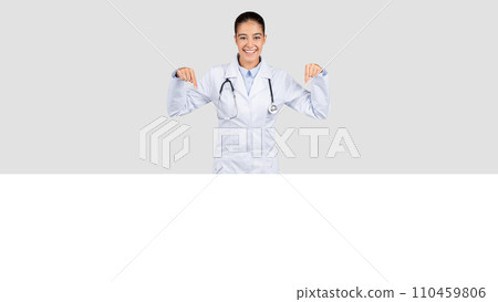Joyful Caucasian millennial doctor with a stethoscope around her neck pointing downwards 110459806