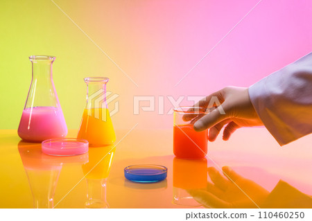 Front view of colorful solution inside erlenmeyer flask, beaker and transparent petri dish on yellow pink gradient background. A hand holding a glassware. Free space for display. 110460250