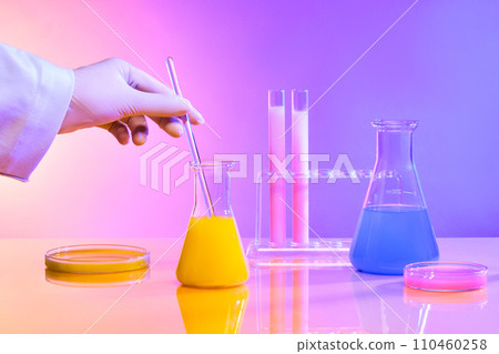 A hand stirs a glass rod in an Erlenmeyer flask with pink liquid, beside glassware holding colorful liquids. The backdrop transitions from pink to purple in this close-up scene. 110460258