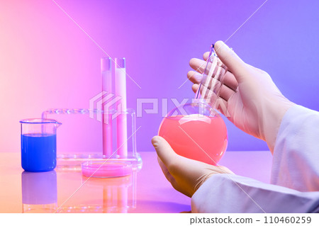 The researcher's hands are holding a boiling flask containing a pink solution, next to glassware containing brightly colored liquids. Pink background turns purple. 110460259