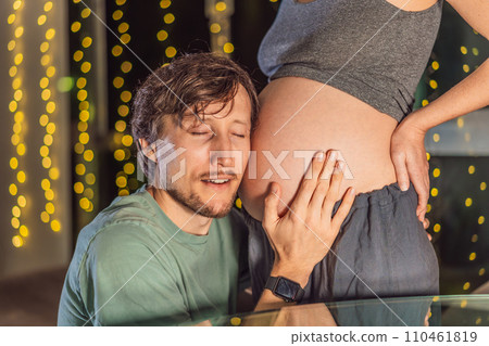 In a tender holiday moment, a husband kisses his wife's pregnant belly, expressing love and anticipation for the Christmas joy to come In a tender holiday moment, a husband kisses his wife's pregnant belly, expressing love and anticipation for the Christmas joy to come 110461819