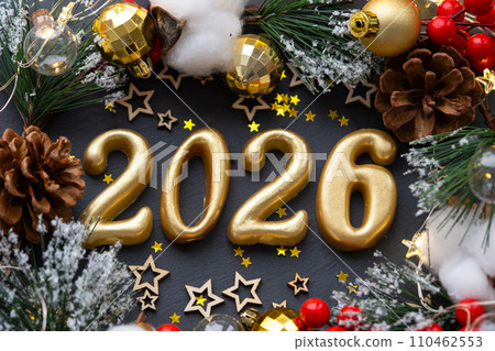 The golden figures 2026 made of candles on a black stone slate background are decorated with a festive decor of stars, sequins, fir branches, balls and garlands. Greeting card, happy New Year. The golden figures 2026 made of candles on a black stone slate background are decorated with a festive decor of stars, sequins, fir branches, balls and garlands. Greeting card, happy New Year. 110462553