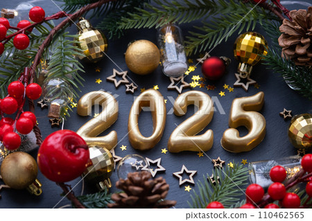 The golden figures 2025 made of candles on a black stone slate background are decorated with a festive decor of stars, sequins, fir branches, balls and garlands. Greeting card, happy New Year. The golden figures 2025 made of candles on a black stone slate background are decorated with a festive decor of stars, sequins, fir branches, balls and garlands. Greeting card, happy New Year. 110462565