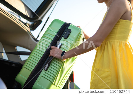 Young woman in summer dress putting green suitcase inside her car trunk. Travel and vacations concept. Young woman in summer dress putting green suitcase inside her car trunk. Travel and vacations concept. 110462744