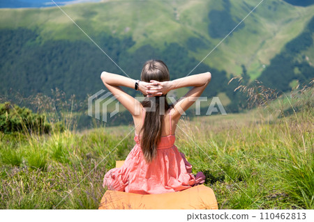 Young woman in red dress resting on green grassy field on a sunny day in summer mountains enjoying view of nature. 110462813
