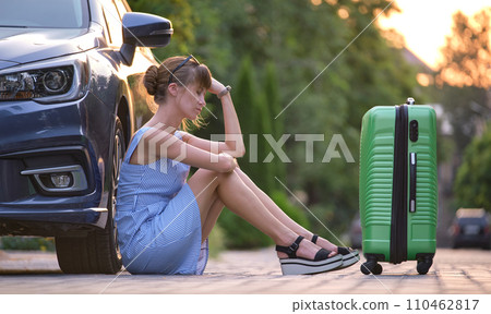 Young tired woman with suitcase sitting beside car waiting for someone. Travelling and vacations concept. 110462817