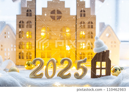 Golden figures number 2025 and tiny home on background of cozy windows of a house with warm light with festive decor of stars,snow and garlands. Greeting card, Happy New Year, cozy home 110462983