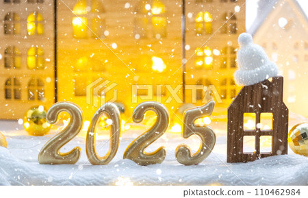 Golden figures number 2025 and tiny home on background of cozy windows of a house with warm light with festive decor of stars,snow and garlands. Greeting card, Happy New Year, cozy home 110462984