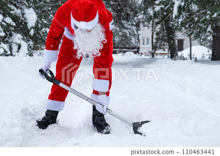 Santa Claus cleans snow with shovel in winter outdoors after a snowfall. Cleaning the streets in the village, clearing the passage for cars, difficult weather conditions for Christmas and New Year 110463441