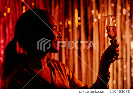 Side view of cheerful girl in red dim light having fun at dance club party while looking at glass of champagne in raised hand Side view of cheerful girl in red dim light having fun at dance club party while looking at glass of champagne in raised hand 110463678