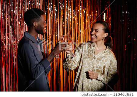 Waist up shot of two cheerful friends at club party toasting with glasses of champagne while standing at red foil fringe curtains Waist up shot of two cheerful friends at club party toasting with glasses of champagne while standing at red foil fringe curtains 110463681