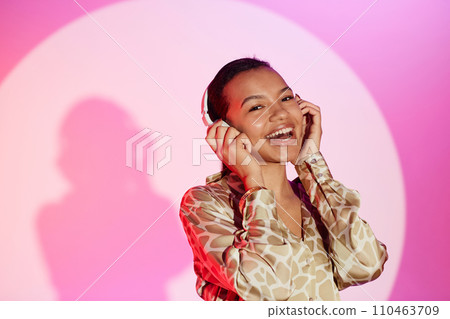Medium shot of laughing young woman standing at bright pink background looking at camera with hand on headphones listening to upbeat music Medium shot of laughing young woman standing at bright pink background looking at camera with hand on headphones listening to upbeat music 110463709
