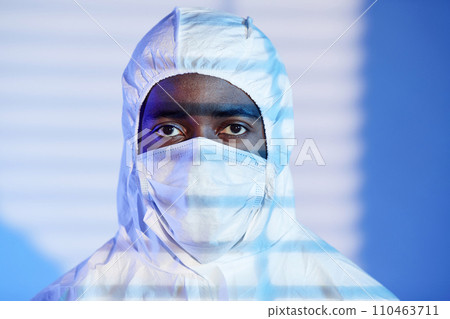 Close up shot of African American scientist looking at camera dressed in white hazmat suit and mask while standing in blue light and blinds shadow 110463711