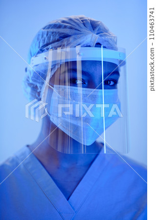 Headshot of female medic wearing protective face shield with sterile hat and medical mask standing in studio with blue neon light and looking at camera 110463741