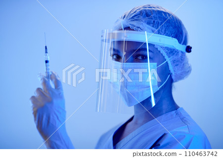 Medium close up shot of woman doctor in medical protective wear holding syringe while looking at camera in blue neon light 110463742