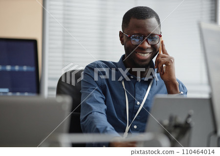Waist up shot of cheerful Black man wearing glasses and blue shirt sitting in IT office while talking to customer using smartphone 110464014