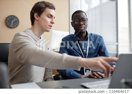Waist up shot of young Caucasian male programmer pointing at laptop screen and talking to African American coworker while working in office Waist up shot of young Caucasian male programmer pointing at laptop screen and talking to African American coworker while working in office 110464017