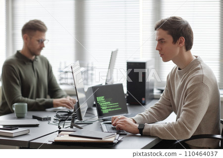 Side view of young Caucasian male IT specialist looking at computer screen and typing on keyboard while sitting at desk with coworker in blurred background 110464037