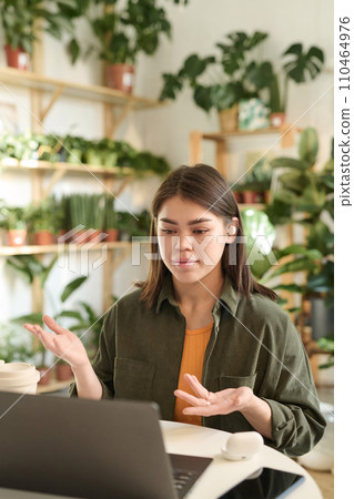 Medium shot of young plant store worker having business call via her laptop at workplace 110464976