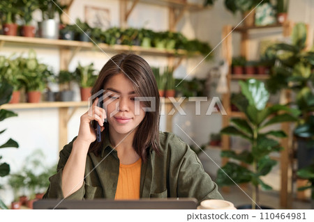 Young plant store manager talking on mobile phone while browsing laptop sitting at desk at store 110464981