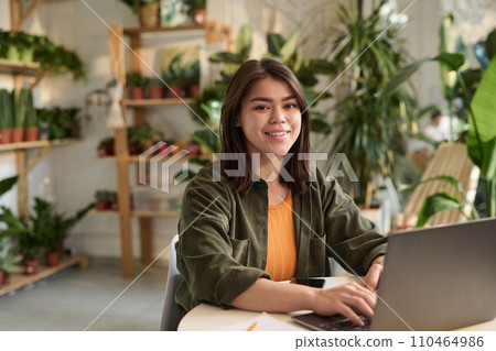 Medium shot of young female plant store owner sitting at her workplace with laptop smiling at camera Medium shot of young female plant store owner sitting at her workplace with laptop smiling at camera 110464986