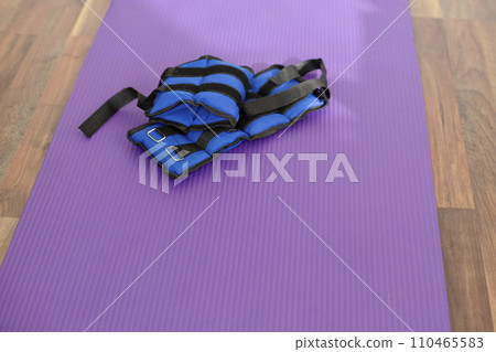 Fabric blue leg weights lie on a yoga mat. Sport equipment. Purple yoga mat. Fabric blue leg weights lie on a yoga mat. Sport equipment. Purple yoga mat. 110465583