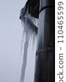 A huge ice icicle hangs on the edge of the roof of a multi-story building. An icicle hangs near a metal pipe for ventilation. 110465599