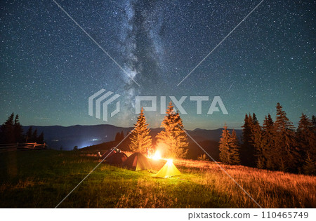 Night camping in mountains under starry sky. Tourist tents in campsite under beautiful sky full of stars with Milky way. Silhouette of hikers having a rest near burning campfire. Concept of tourism. Night camping in mountains under starry sky. Tourist tents in campsite under beautiful sky full of stars with Milky way. Silhouette of hikers having a rest near burning campfire. Concept of tourism. 110465749