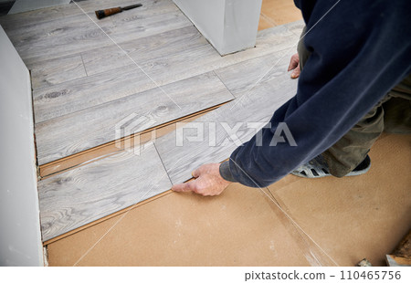 Close up of man construction worker installing laminate timber flooring in apartment under renovation. Male hands placing laminate planks on the floor at home. Flooring renovation concept. Close up of man construction worker installing laminate timber flooring in apartment under renovation. Male hands placing laminate planks on the floor at home. Flooring renovation concept. 110465756