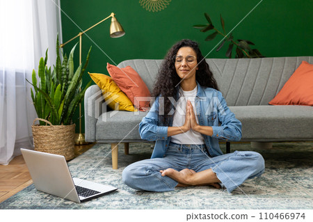 Young woman resting meditating in lotus pose sitting on floor at home in living room cinema, Hispanic woman with laptop doing online group yoga in training class remotely. 110466974