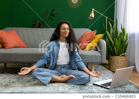 Young woman resting meditating in lotus pose sitting on floor at home in living room cinema, Hispanic woman with laptop doing online group yoga in training class remotely. Young woman resting meditating in lotus pose sitting on floor at home in living room cinema, Hispanic woman with laptop doing online group yoga in training class remotely. 110466983