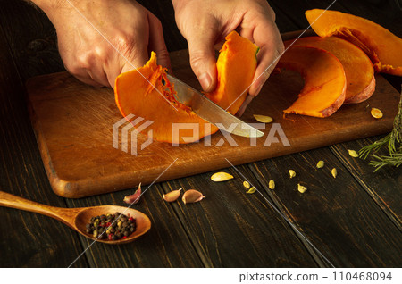 The chef's hand uses a knife to cut a slice of pumpkin on the kitchen board. The concept of preparing ingredients for a delicious pie The chef's hand uses a knife to cut a slice of pumpkin on the kitchen board. The concept of preparing ingredients for a delicious pie 110468094