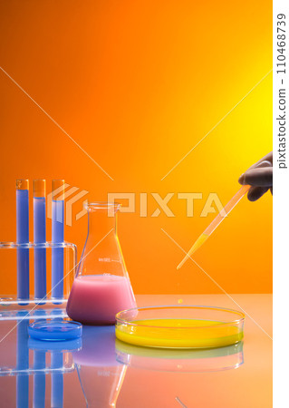 Over the gradient background, hand model with white glove is dripping yellow chemical into a large petri dish. Chemistry lab laboratory technical equipment with many glassware bottle 110468739