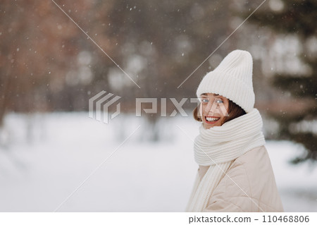 Happy smiling young woman portrait dressed a coat scarf and a hat and the mittens enjoys winter weather at the snowy winter park Happy smiling young woman portrait dressed a coat scarf and a hat and the mittens enjoys winter weather at the snowy winter park 110468806