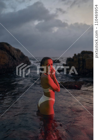 Attractive female enjoys serene night dip in ocean illuminated by crimson glow. Travel enthusiast indulges in after-dark swimming, ocean waves gently lapping around. Beauty of nocturnal water. 110469954